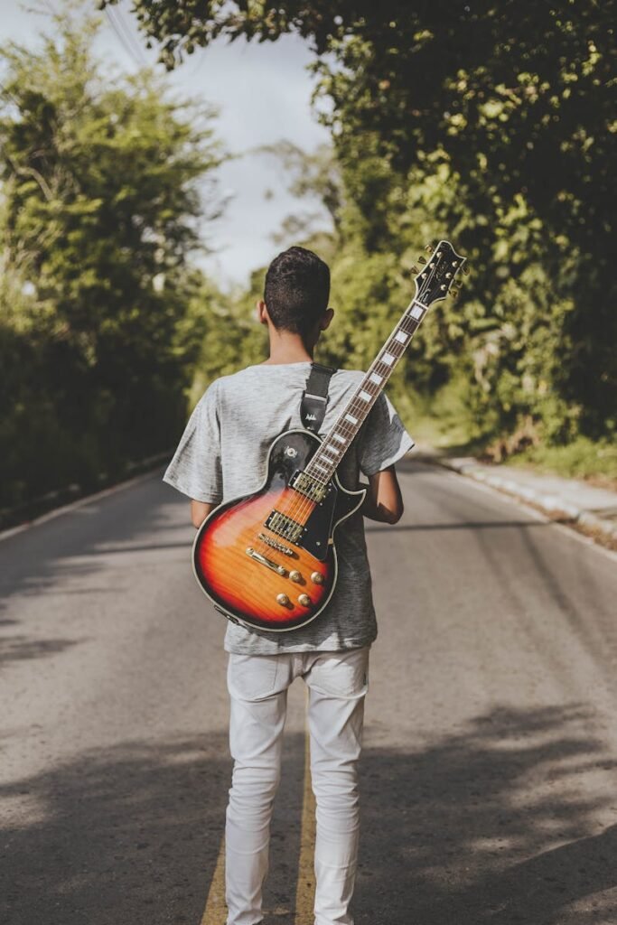 A young man carrying an electric guitar on his back walks down a tree-lined road.
